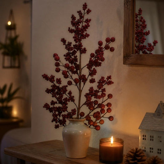 Vara Fruto de Oso 73 cm Roja — ramas de bayas rojas en un florero de cerámica junto a una vela encendida